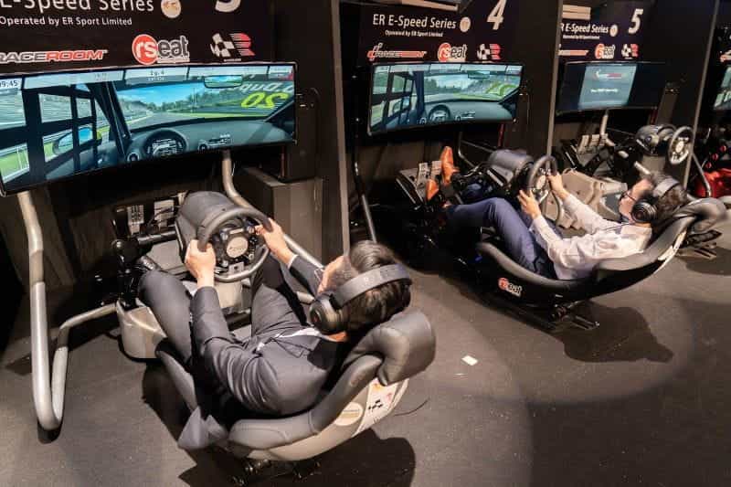 A pair of esports gamers playing a racing game while sitting in racing chairs and using steering wheels.