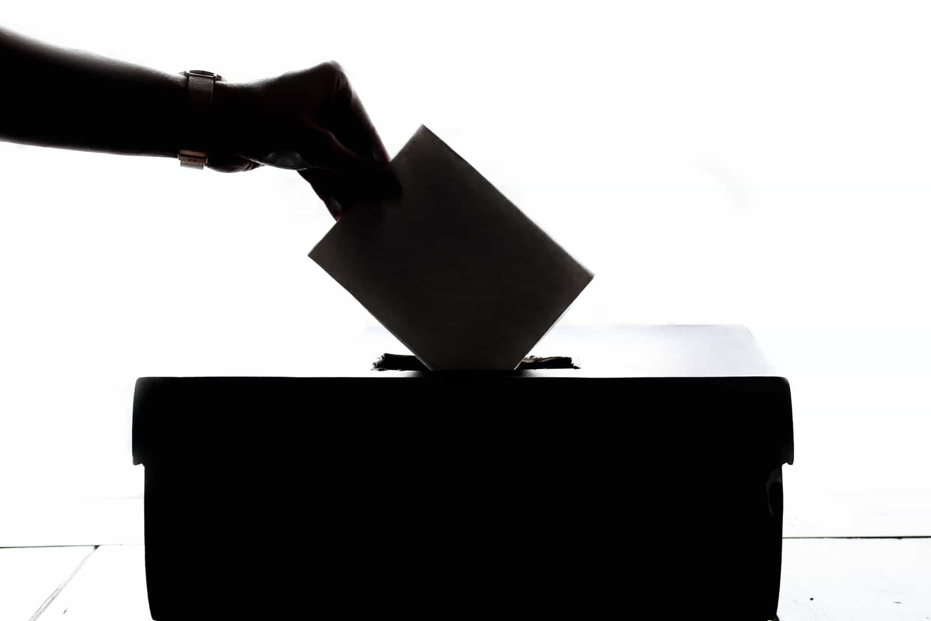 A backlit, silhouetted hand drops a ballot into a ballot box.